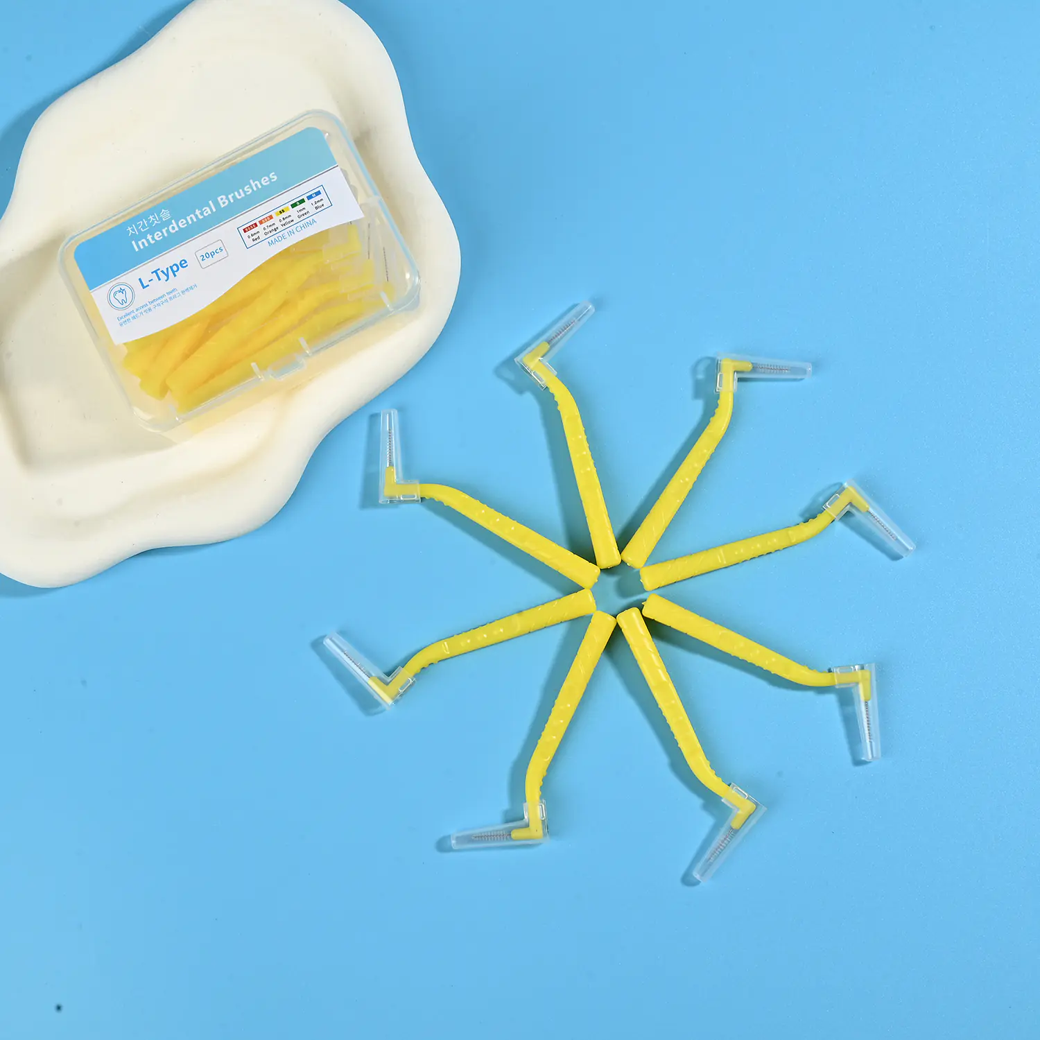 Yellow L-type interdental cleaners arranged radial star shape alongside original clear plastic packaging box on irregular beige pad and bright blue background